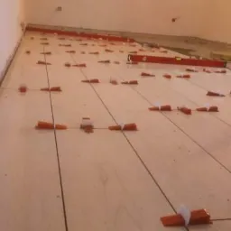 Układanie jasnych płytek podłogowych z użyciem systemu poziomowania, widoczne pomarańczowe kliny i czarne krzyżyki dystansowe.