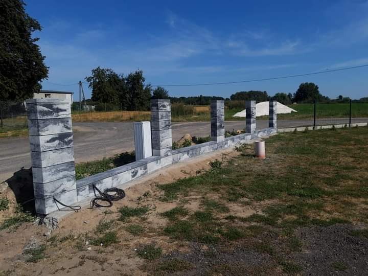 Wznoszona brama wjazdowa z szarej kostki betonowej, widoczne słupki i podmurówka w trakcie budowy, zwinięty kabel na ziemi, w tle pola i drzewa.