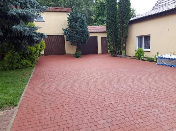 Czerwona kostka brukowa na podjeździe do garażu w domu jednorodzinnym, z widocznym trawnikiem i iglakami po bokach.
