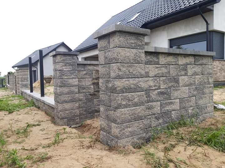 Nowoczesny murek oporowy z szarej kostki łupanej, zakończony filarami, na tle domu z ciemną dachówką, widoczne ślady prac ziemnych.