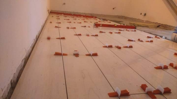 Układanie jasnych płytek podłogowych z użyciem systemu poziomowania, widoczne pomarańczowe kliny i czarne krzyżyki dystansowe.