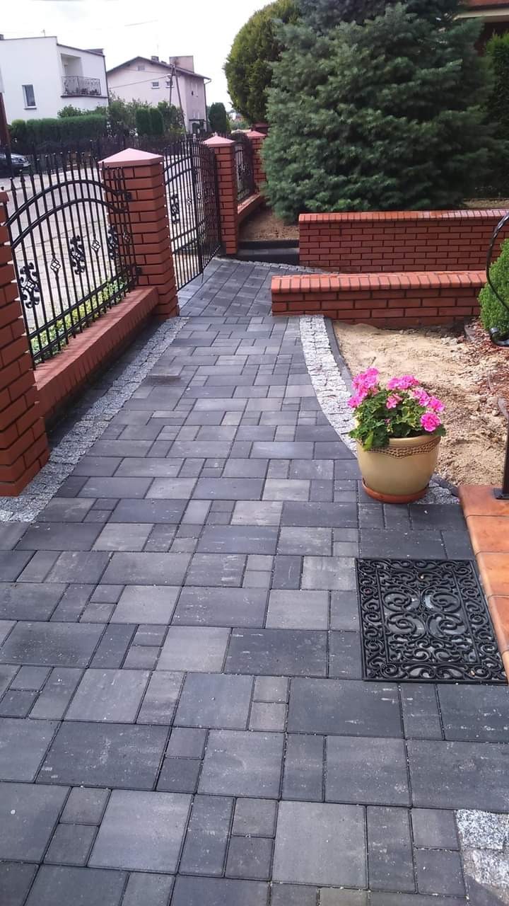 Ułożona kostka brukowa w odcieniach szarości tworząca ścieżkę z obramowaniem z drobnych kamieni, ograniczona ceglanymi słupkami z czarnym, kutym ogrodzeniem, donica z różowymi kwiatami...