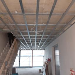 ELB-BUD - Montaż metalowego stelażu sufitu podwieszanego z widocznymi przewodami elektrycznymi, fragment schodów i okna w tle.