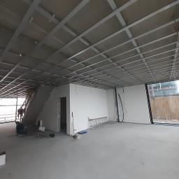 ELB-BUD - Montaż metalowego rusztu sufitowego w przestronnym pomieszczeniu z dużymi oknami, widoczna drabina i częściowo obrobione ściany, w tle schody i pracownik.