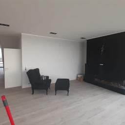 ELB-BUD - Jasne, przestronne wnętrze z drewnianą podłogą, czarnym kominkiem i fotelami, widok na zewnątrz przez duże okna, częściowo widoczny stół z krzesłami.