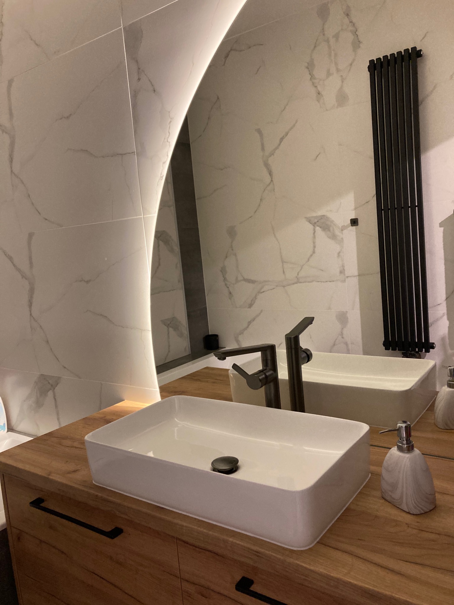 Nowoczesna łazienka z umywalką nablatową, baterią w kolorze grafitowym, podświetlanym lustrem LED i grzejnikiem drabinkowym na ścianie wyłożonej płytkami imitującymi marmur.
