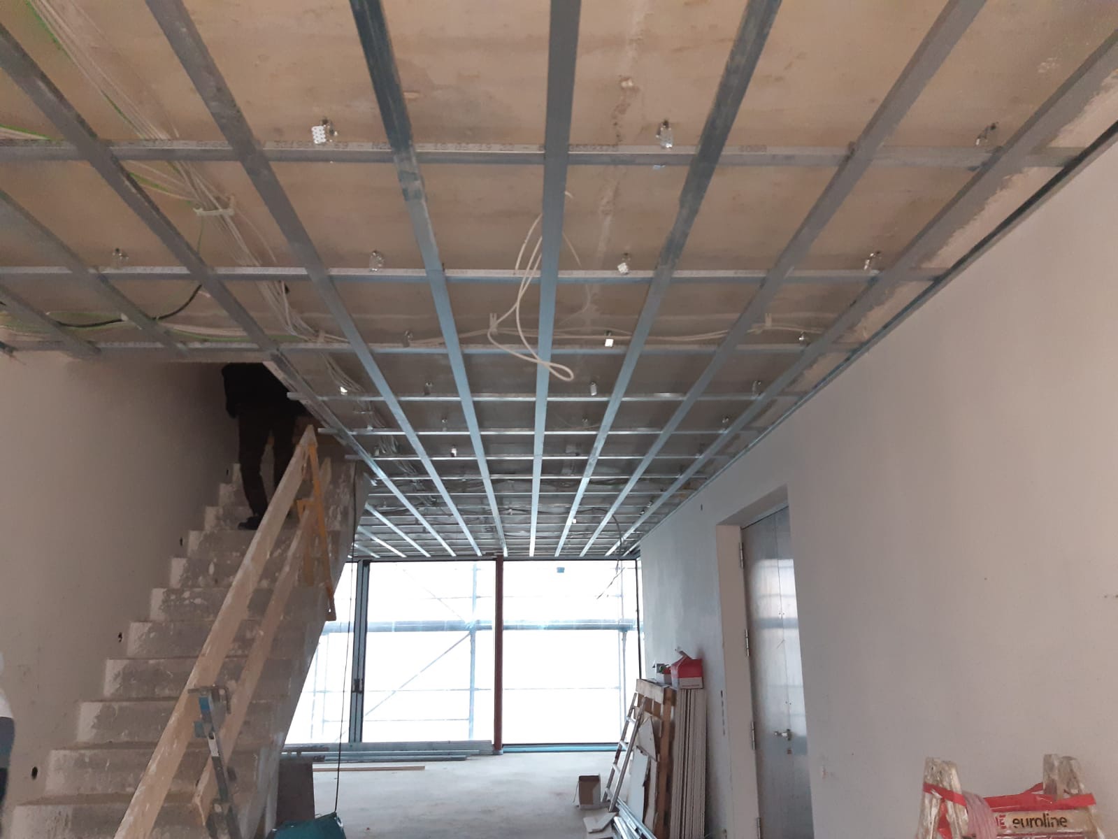 Montaż metalowego stelażu sufitu podwieszanego z widocznymi przewodami elektrycznymi, fragment schodów i okna w tle.