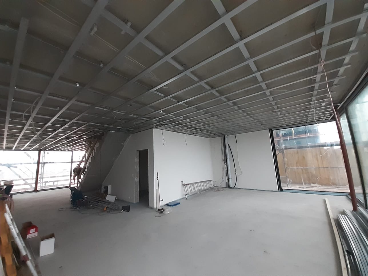 Montaż metalowego rusztu sufitowego w przestronnym pomieszczeniu z dużymi oknami, widoczna drabina i częściowo obrobione ściany, w tle schody i pracownik.