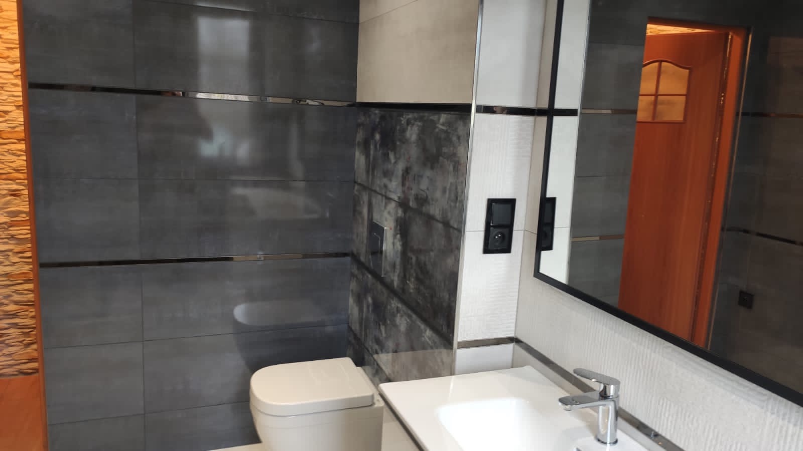 Nowoczesna łazienka z szarymi płytkami wielkoformatowymi z metalowymi listwami, imitacją kamienia, białą ceramiką i lustrem z czarną ramą.