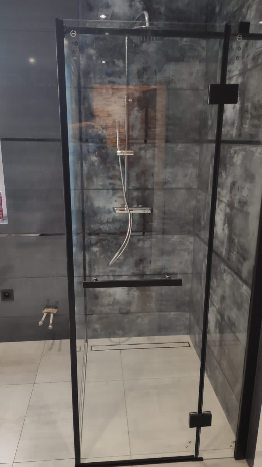 Nowoczesna kabina prysznicowa z czarnymi profilami, szklane ściany, deszczownica i bateria termostatyczna na tle szarej ściany wykończonej płytkami imitującymi beton.