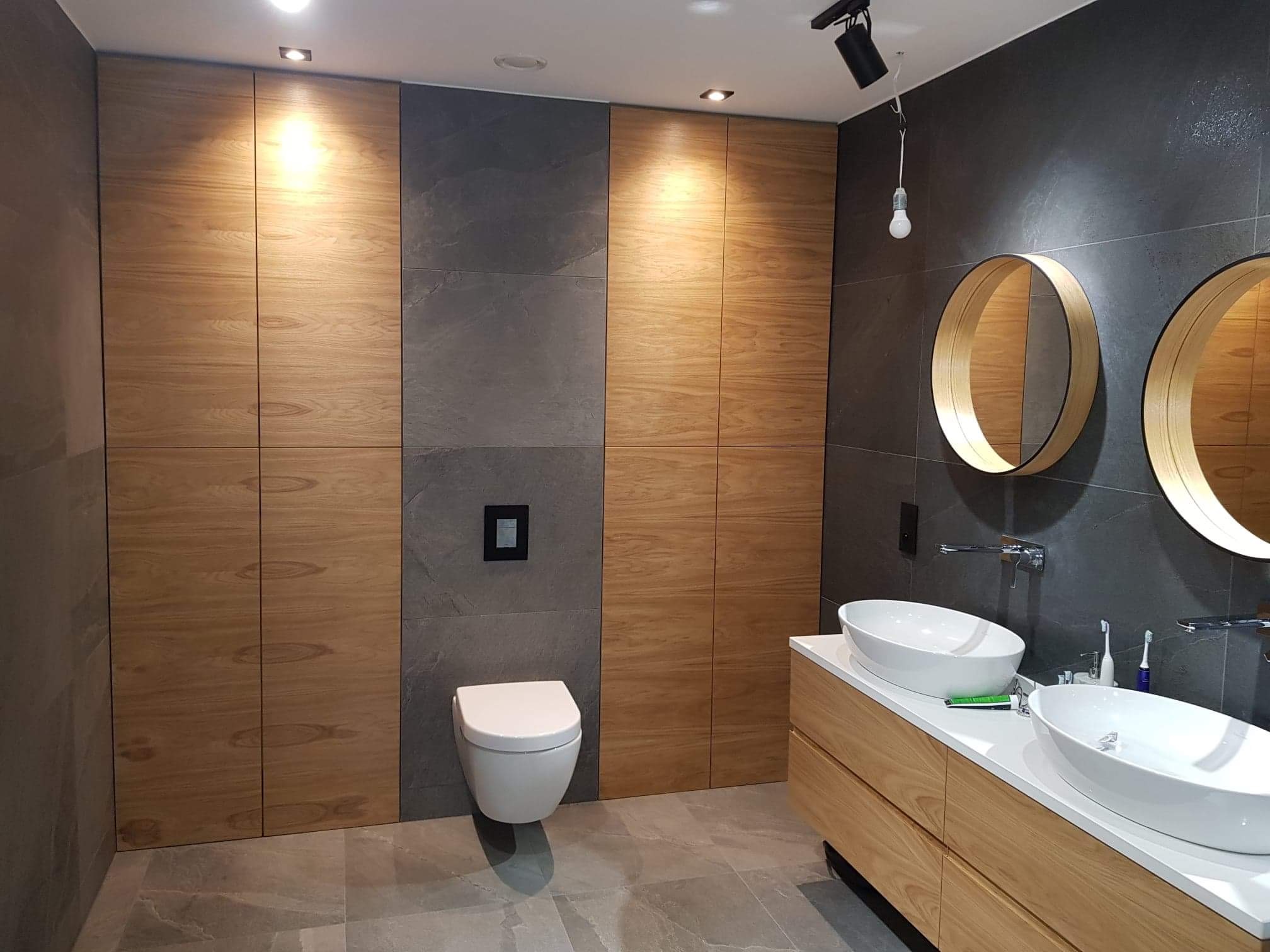 Nowoczesna łazienka z drewnianymi szafkami, szarymi płytkami, okrągłymi lustrami i podwieszaną toaletą. Widoczna surowa żarówka na kablu.