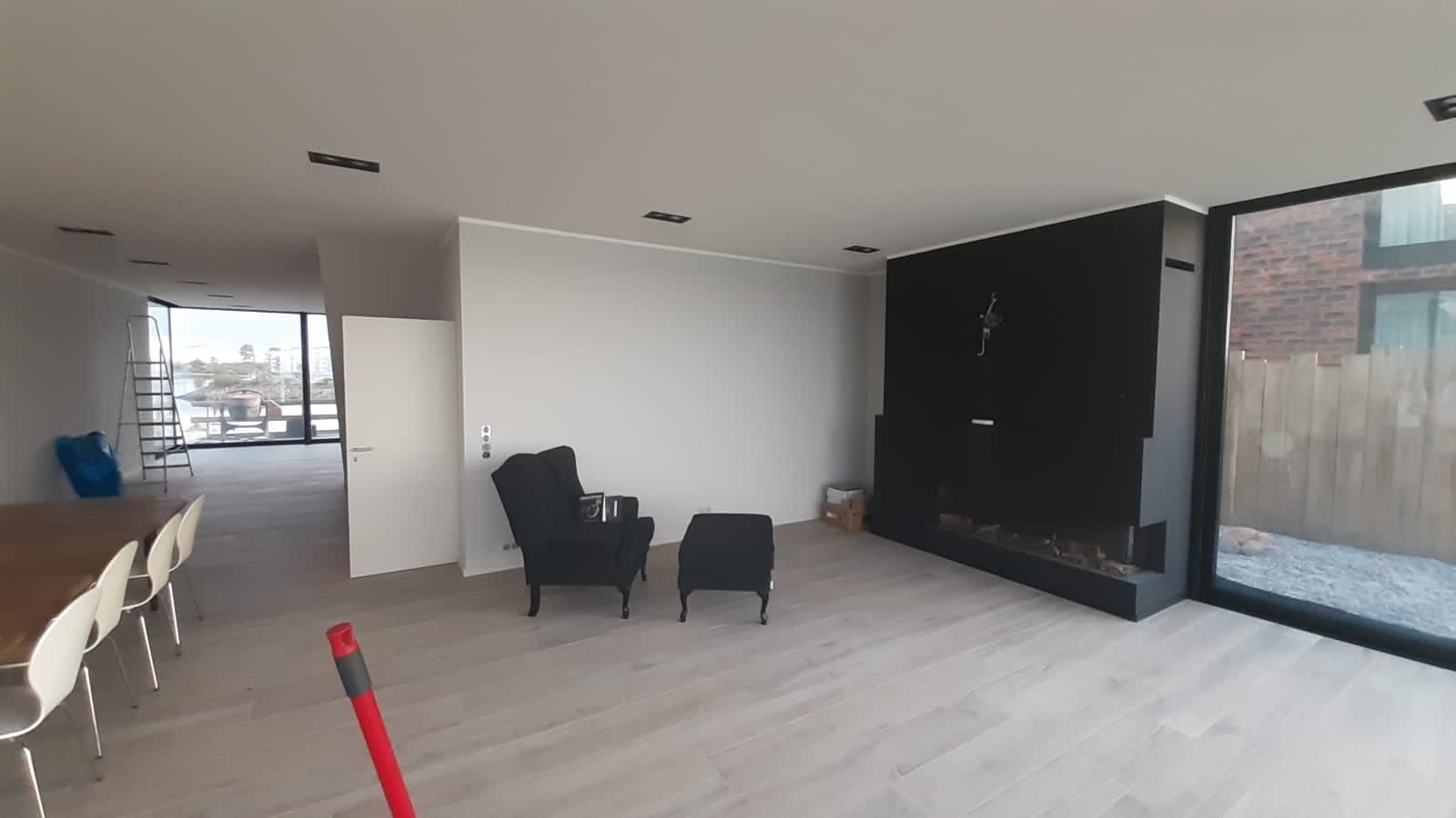 Jasne, przestronne wnętrze z drewnianą podłogą, czarnym kominkiem i fotelami, widok na zewnątrz przez duże okna, częściowo widoczny stół z krzesłami.