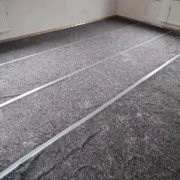 Przygotowanie podłoża pod ogrzewanie podłogowe: izolacja z wełny mineralnej z aluminiowymi profilami mocującymi, widok z góry na surowy stan pomieszczenia z grzejnikami na ścianach.