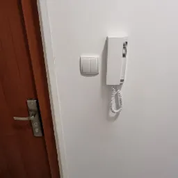 Biały domofon, włącznik światła i gniazdko elektryczne zamontowane na białej ścianie obok brązowych drzwi wejściowych z metalową klamką, widoczne szare płytki podłogowe.