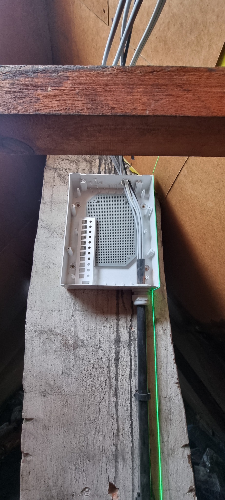 Otwarta puszka elektryczna z wyprowadzonymi przewodami, zamontowana na betonowym słupie w surowym wnętrzu, z widoczną wiązką kabli i zieloną linią lasera wyznaczającą pion.