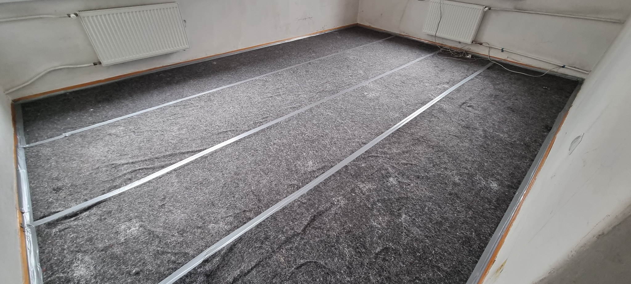 Przygotowanie podłoża pod ogrzewanie podłogowe: izolacja z wełny mineralnej z aluminiowymi profilami mocującymi, widok z góry na surowy stan pomieszczenia z grzejnikami na ścianach.