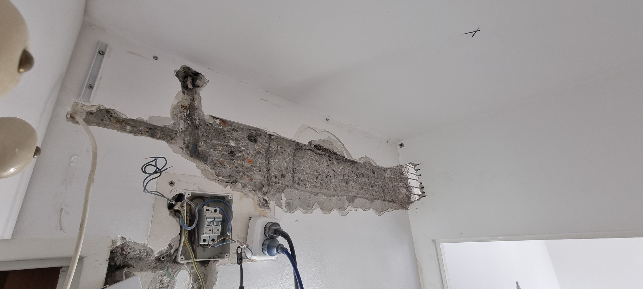 Uszkodzona ściana z odsłoniętym zbrojeniem i instalacją elektryczną w mieszkaniu we Wrocławiu, widoczne kable i gniazdka.