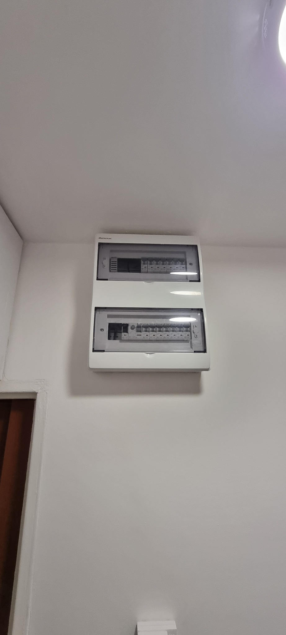 Dwu-segmentowa, biała rozdzielnica elektryczna zamontowana na świeżo pomalowanej, białej ścianie; widoczne zabezpieczenia wewnątrz obudowy z przezroczystymi drzwiczkami.