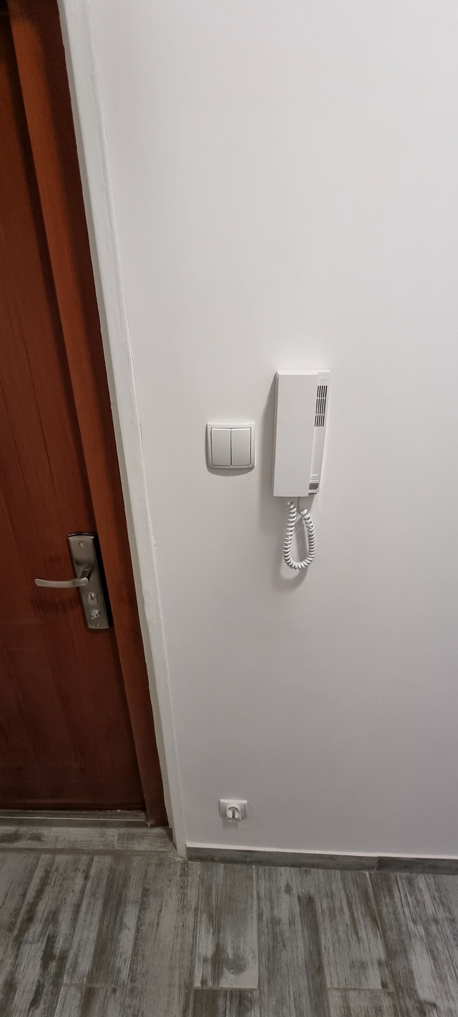 Biały domofon, włącznik światła i gniazdko elektryczne zamontowane na białej ścianie obok brązowych drzwi wejściowych z metalową klamką, widoczne szare płytki podłogowe.