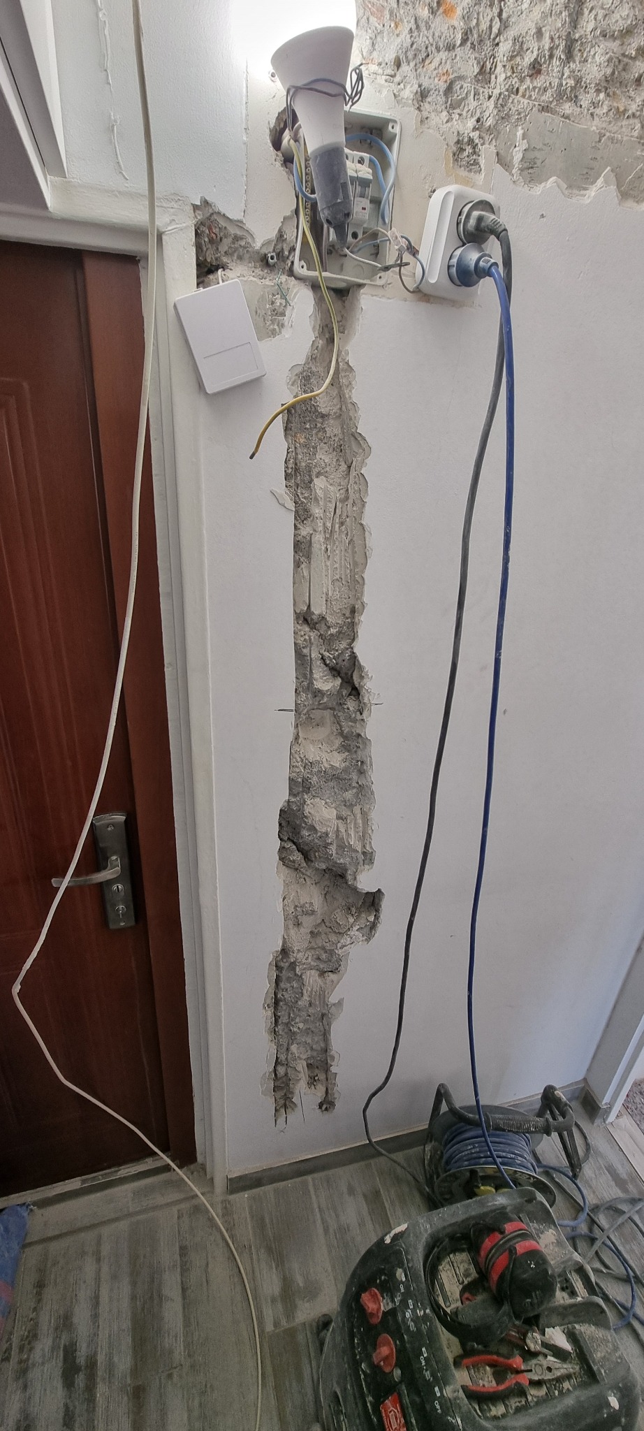 Wykuta bruzda w ścianie obok brązowych drzwi, widoczna puszka elektryczna z przewodami, gniazdo z wpiętymi kablami oraz narzędzia na podłodze w trakcie prac instalacyjnych.