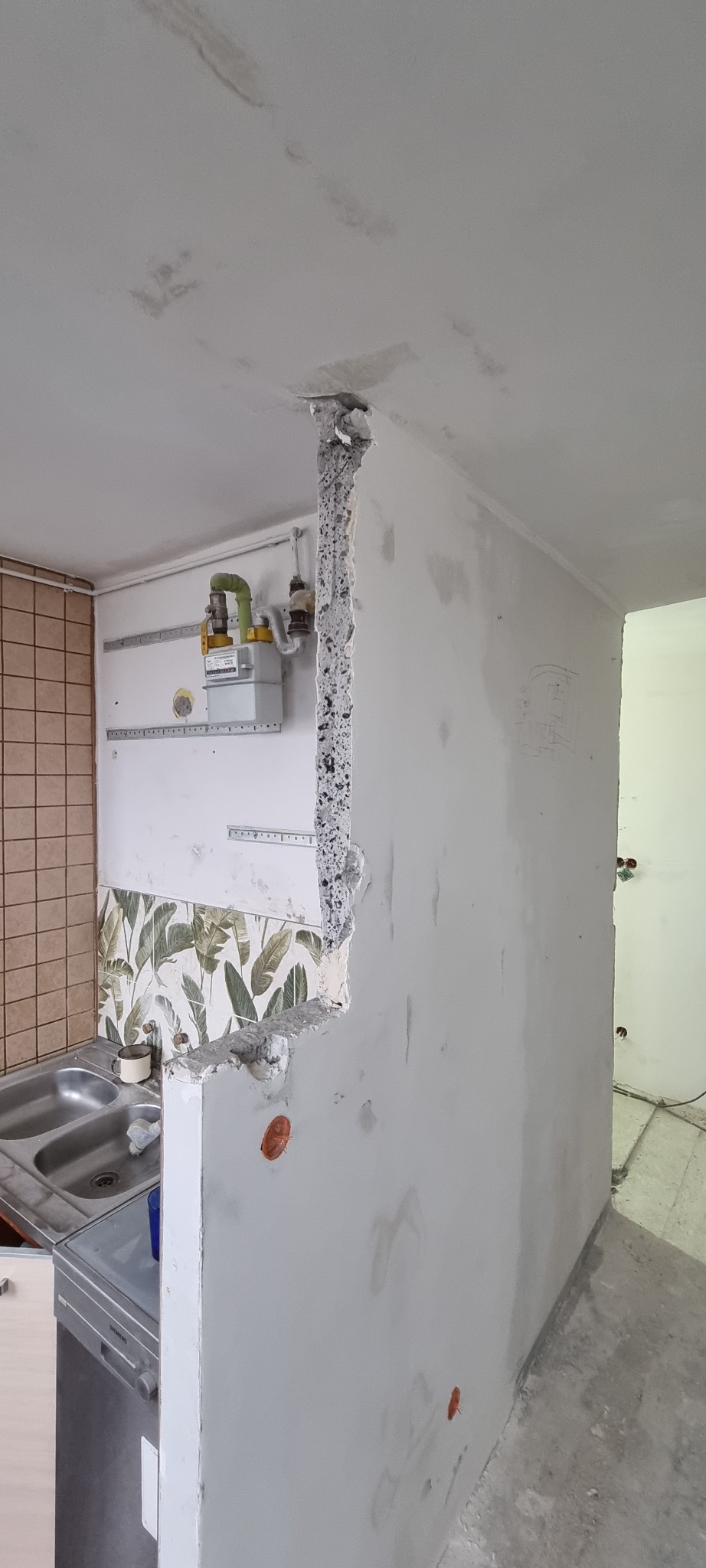 Ściana w trakcie remontu kuchni z widocznym pionowym bruzdowaniem pod instalację elektryczną, obok licznik gazu i fragment zlewu.