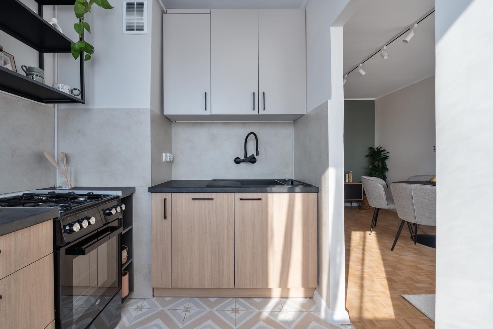 Nowoczesna kuchnia z drewnianymi szafkami, czarnym blatem i armaturą. Widoczny fragment salonu z parkietem i szarymi krzesłami. Oświetlenie punktowe.