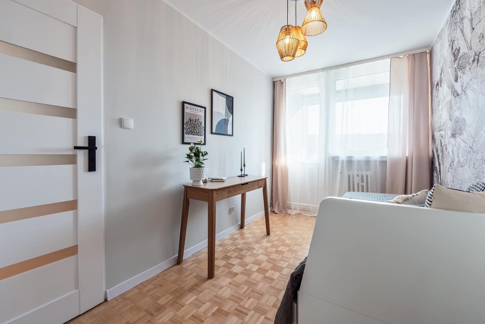 Nowoczesne oświetlenie w sypialni: trzy plecione lampy wiszące nad drewnianą konsolą z dekoracjami, białe drzwi z czarną klamką i wzorzysta tapeta w tle.