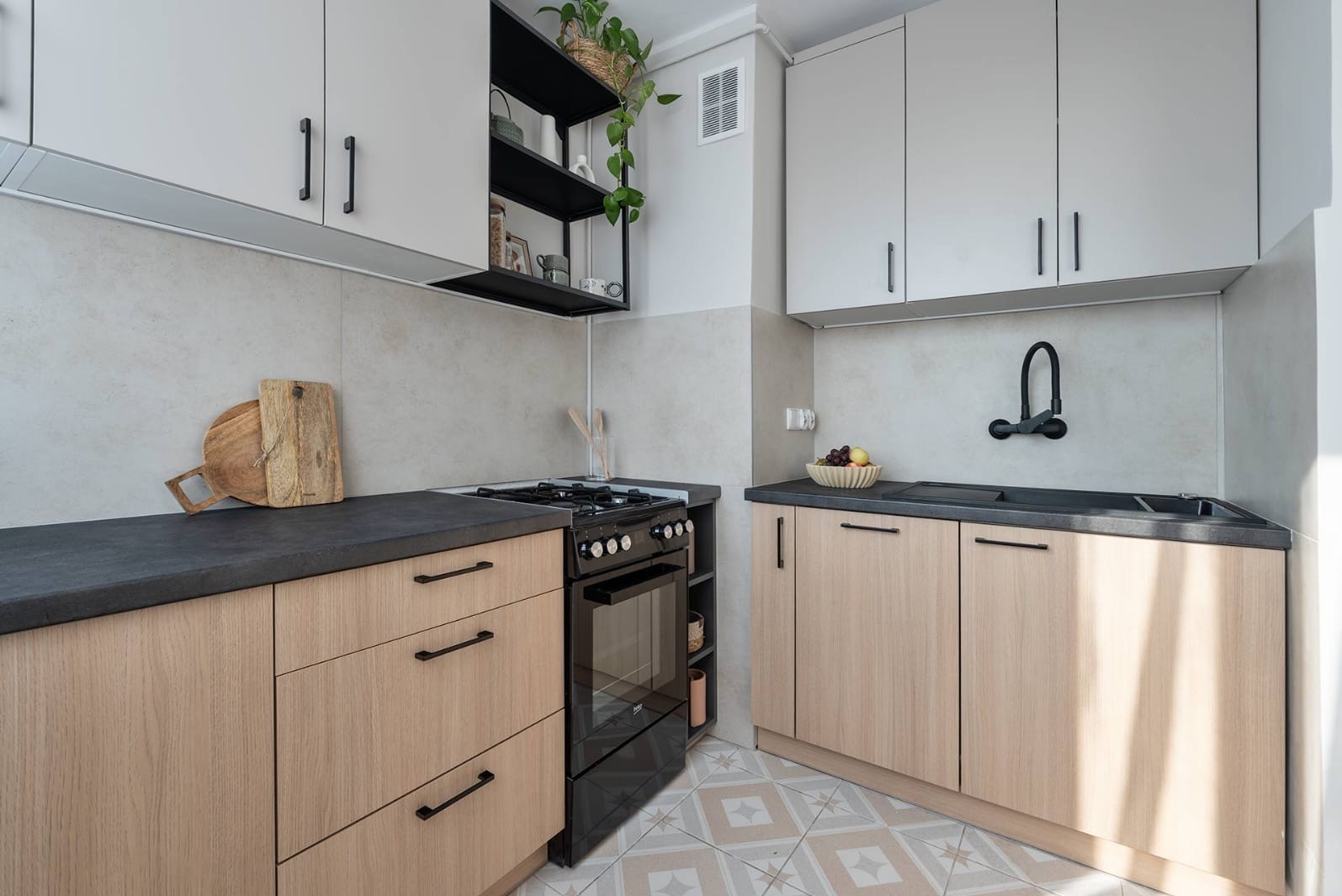 Nowoczesna kuchnia w jasnych barwach z czarnymi akcentami: armatura, piekarnik i półki. Uwagę przykuwają geometryczne płytki podłogowe i drewniane blaty.