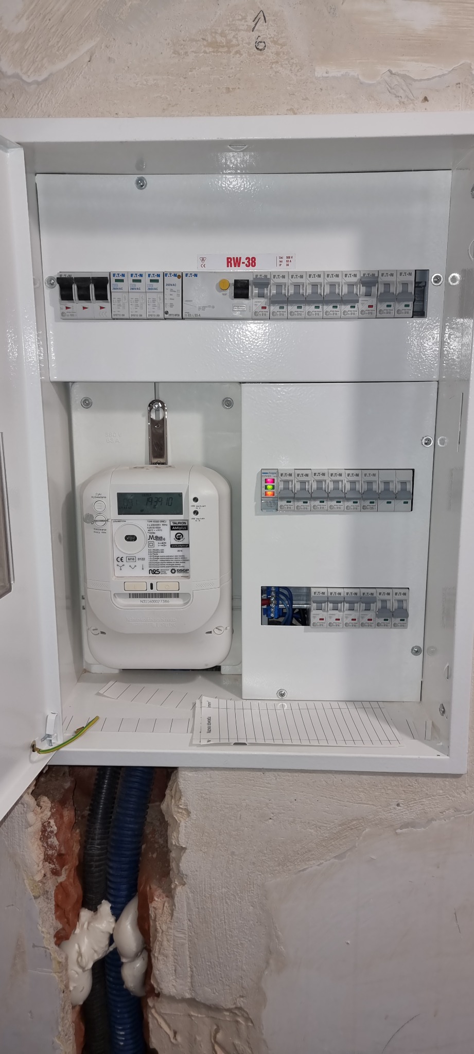 Otwarta skrzynka elektryczna z licznikiem i bezpiecznikami, widoczne kable w ścianie. Biała obudowa, szare ściany. Widok z bliska na instalację elektryczną wewnątrz budynku.