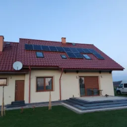 Dom jednorodzinny z czerwoną dachówką i panelami słonecznymi na dachu, widoczny z przodu, z satelitą na ścianie i białym vanem w tle.