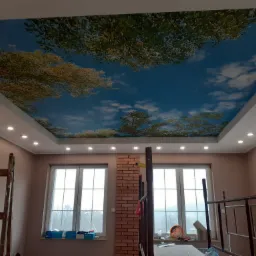 Pokój z zamontowanym sufitem napinanym z motywem nieba i drzew, widoczne drabiny i narzędzia malarskie.