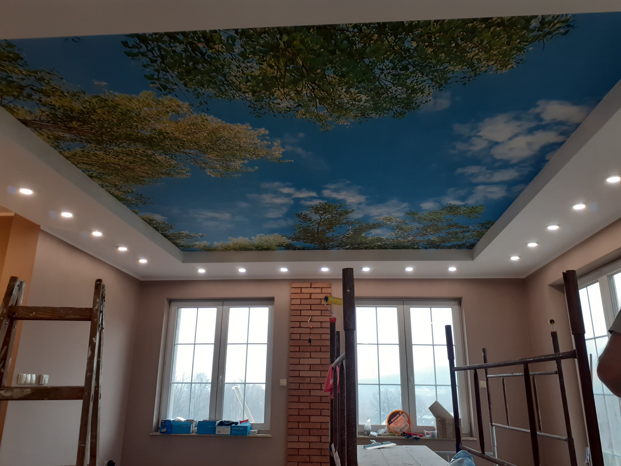 Pokój z zamontowanym sufitem napinanym z motywem nieba i drzew, widoczne drabiny i narzędzia malarskie.
