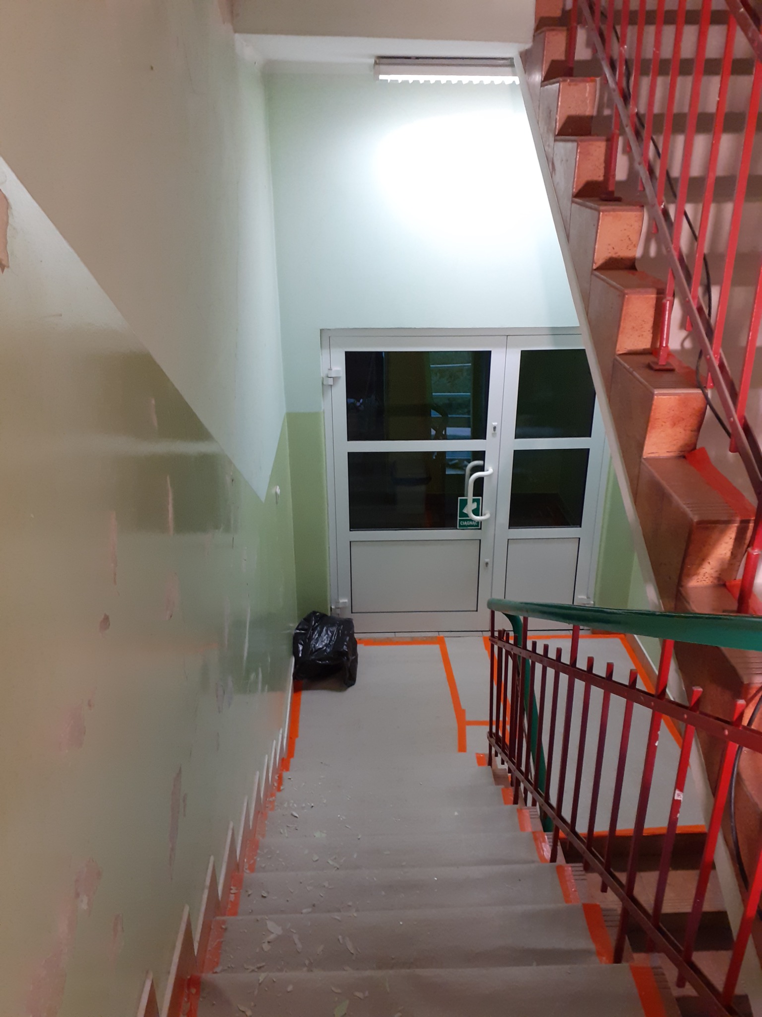 Klatka schodowa w trakcie przygotowania do prac remontowych: zabezpieczone taśmą malarską schody i ściany, widoczne ślady po usuniętej farbie, czarny worek na śmieci przy drzwiach.