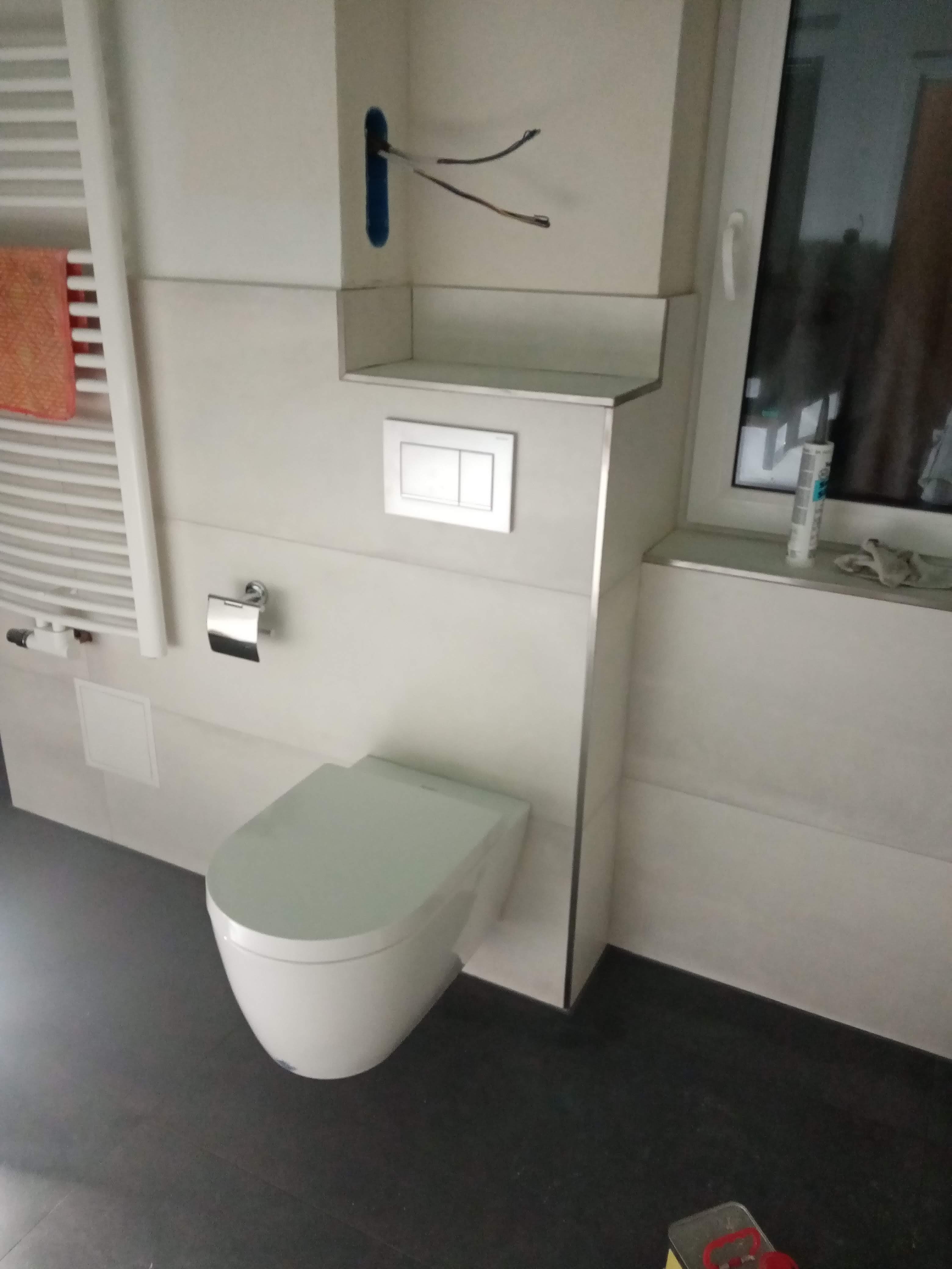 Nowoczesna, podwieszana toaleta w łazience z białymi płytkami, szarą podłogą i grzejnikiem drabinkowym; widoczne wystające przewody elektryczne w ścianie.