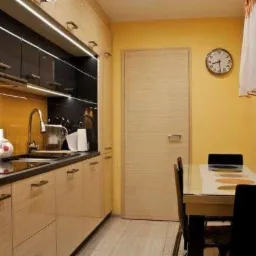 Jasna kuchnia z drewnianymi szafkami, czarnym blatem i żółtą ścianą, widoczny stół z krzesłami i zegar na ścianie.