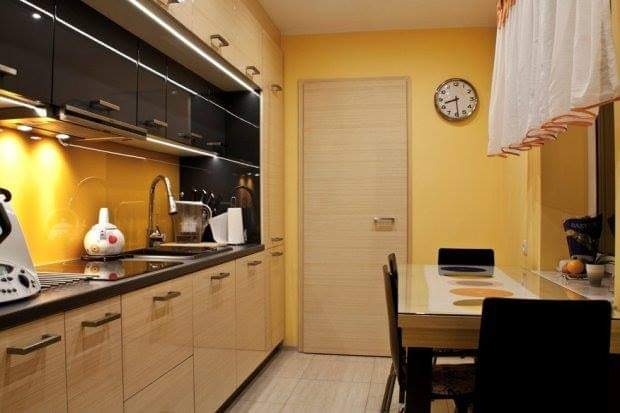 Jasna kuchnia z drewnianymi szafkami, czarnym blatem i żółtą ścianą, widoczny stół z krzesłami i zegar na ścianie.