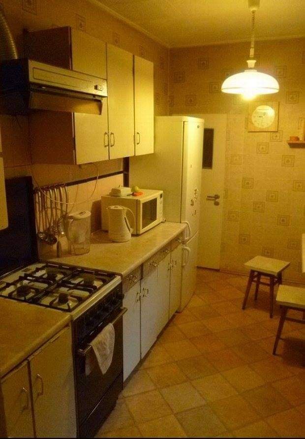 Kuchnia z lat 80-tych z gazową kuchenką, mikrofalówką i białą lodówką, oświetlona pojedynczą lampą wiszącą, z wzorzystą tapetą i dwoma drewnianymi stołkami w tle.