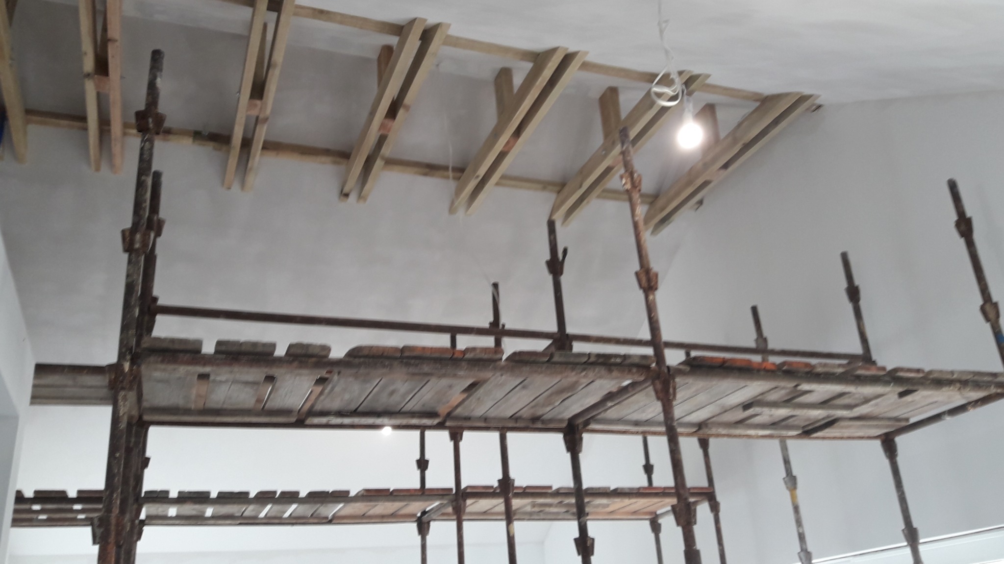 Widok z dołu na rusztowanie w pomieszczeniu z drewnianą konstrukcją pod sufitem i świeżo wykończonymi ścianami w jasnym odcieniu.