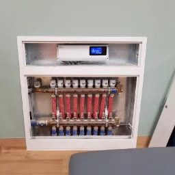 Rozdzielacz ogrzewania podłogowego z widocznymi rurami w kolorze czerwonym i zaworami, umieszczony w białej obudowie, z panelem sterowania na górze, na tle jasnej ściany.