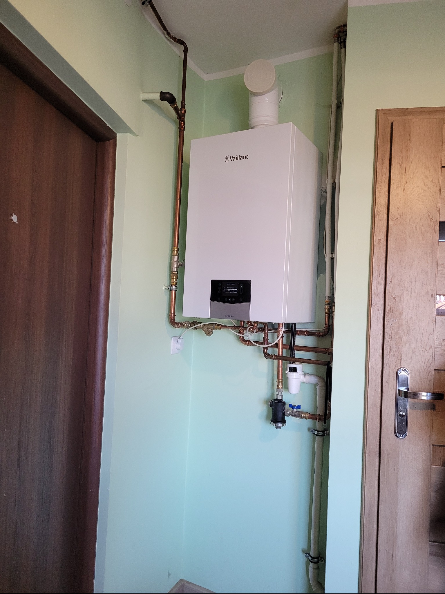 Nowoczesny, biały piec gazowy Vaillant zamontowany w rogu pomieszczenia o miętowych ścianach, z widocznymi miedzianymi rurami i elementami instalacji.