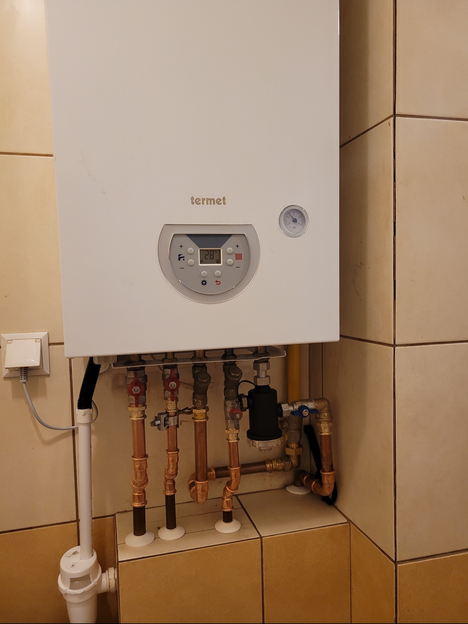 Biały piec gazowy marki Termet zamontowany na kafelkowanej ścianie, widoczne miedziane rury i zawory podłączeniowe, cyfrowy wyświetlacz temperatury na panelu sterowania.