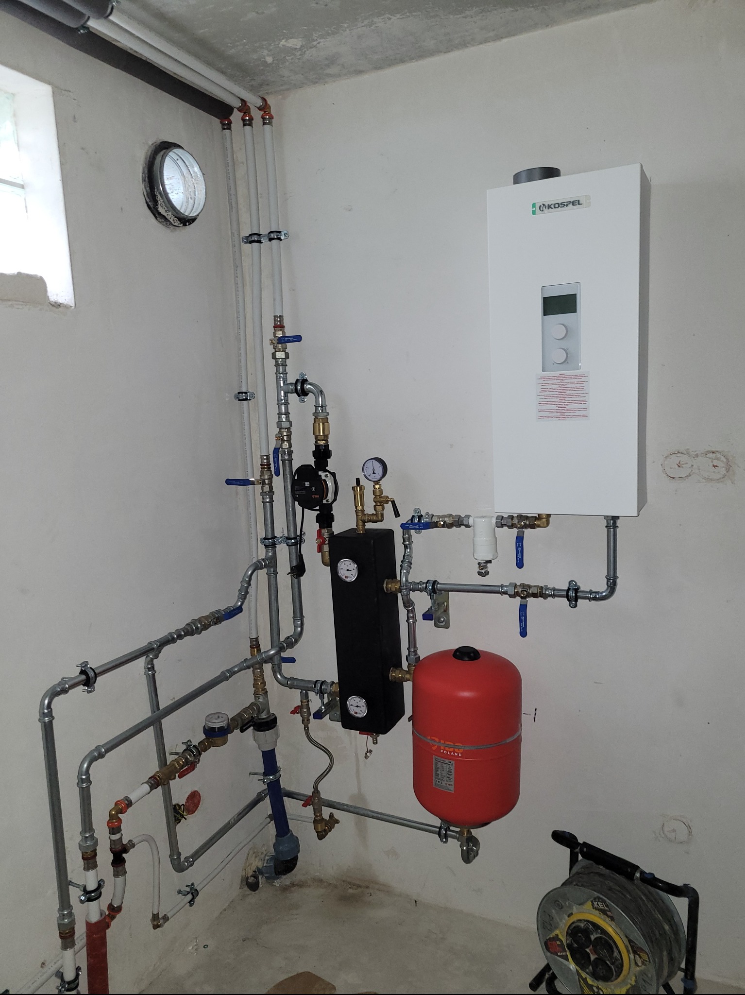 Nowoczesny piec gazowy Kospel zamontowany na ścianie, z widoczną instalacją hydrauliczną, w tym czerwony zbiornik wyrównawczy i liczne rury z zaworami. Na podłodze zwinięty przedłużacz.