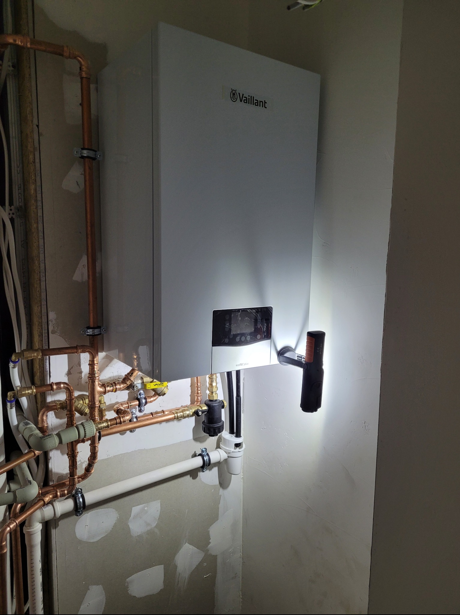Biały piec gazowy Vaillant zamontowany na ścianie, widoczne miedziane rury instalacji, oświetlony latarką magnetyczną.