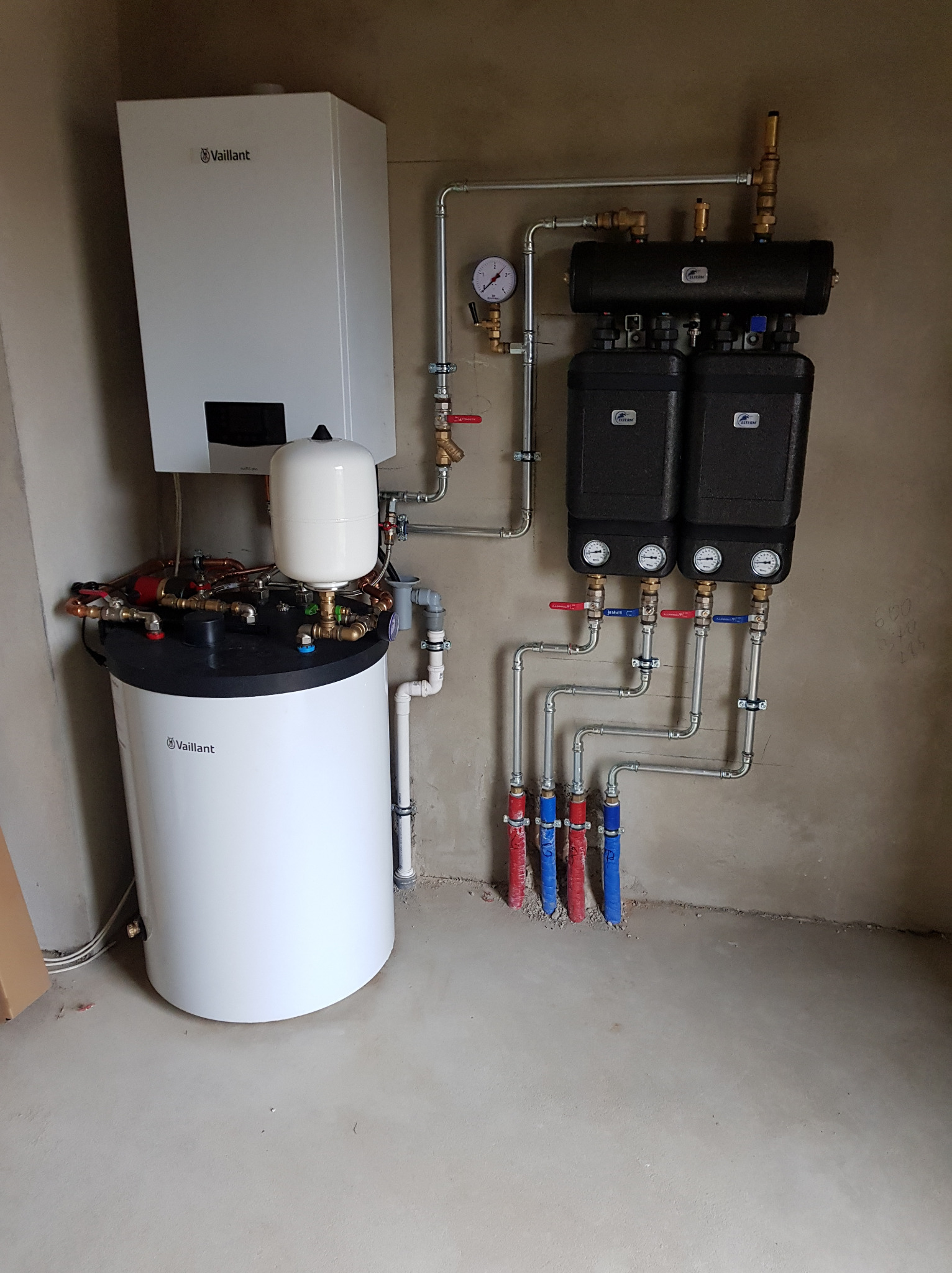 Nowoczesna kotłownia z piecem gazowym Vaillant, zbiornikiem buforowym i rozdzielaczami, rury miedziane i stalowe, manometry, instalacja po montażu, surowy beton na ścianach i posadzce.