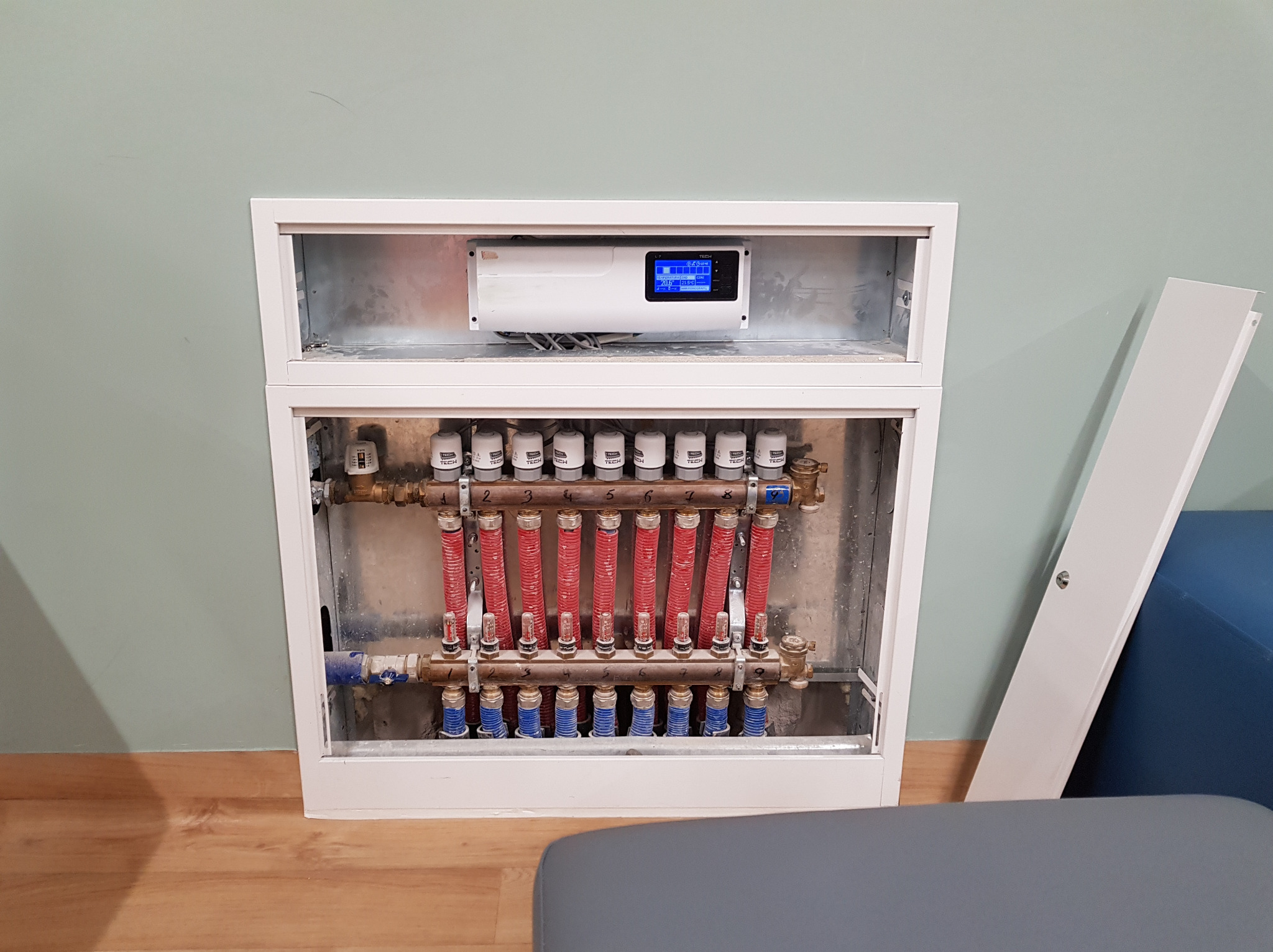 Rozdzielacz ogrzewania podłogowego z widocznymi rurami w kolorze czerwonym i zaworami, umieszczony w białej obudowie, z panelem sterowania na górze, na tle jasnej ściany.