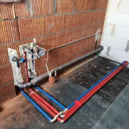 Surowy stan: instalacja wodna z czerwono-niebieskimi rurami PEX na tle ceglanej ściany z widocznymi przyłączami, manometrem i szarą rurą odpływową.