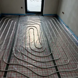 Białe rury ogrzewania podłogowego ułożone na folii z czerwoną kratką, widoczne okno z zimowym krajobrazem na zewnątrz.