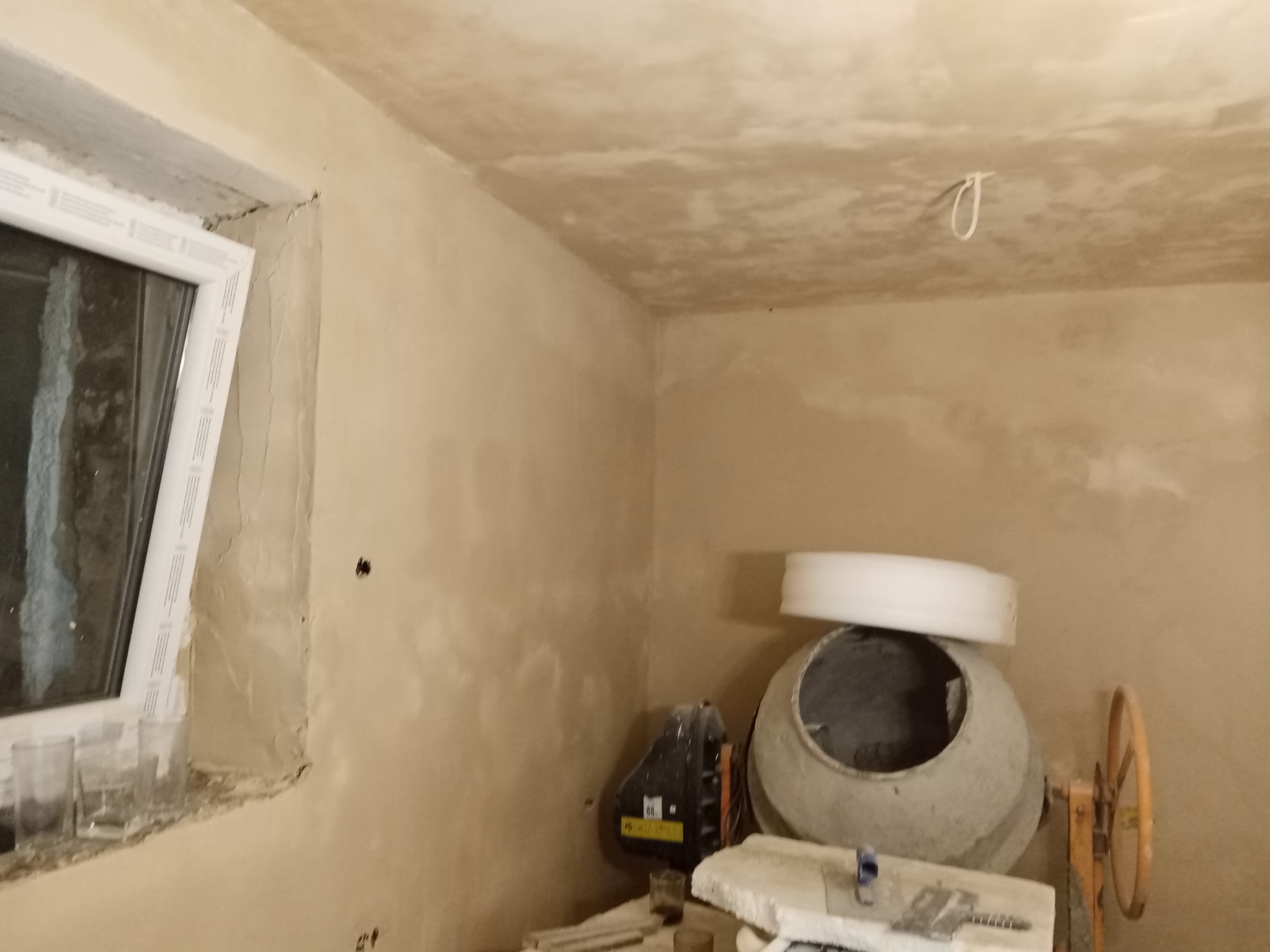Wnętrze w trakcie prac wykończeniowych: ściany i sufit po nałożeniu warstwy tynku, widoczna betoniarka i okno z częściowo obrobionym ościeżem.
