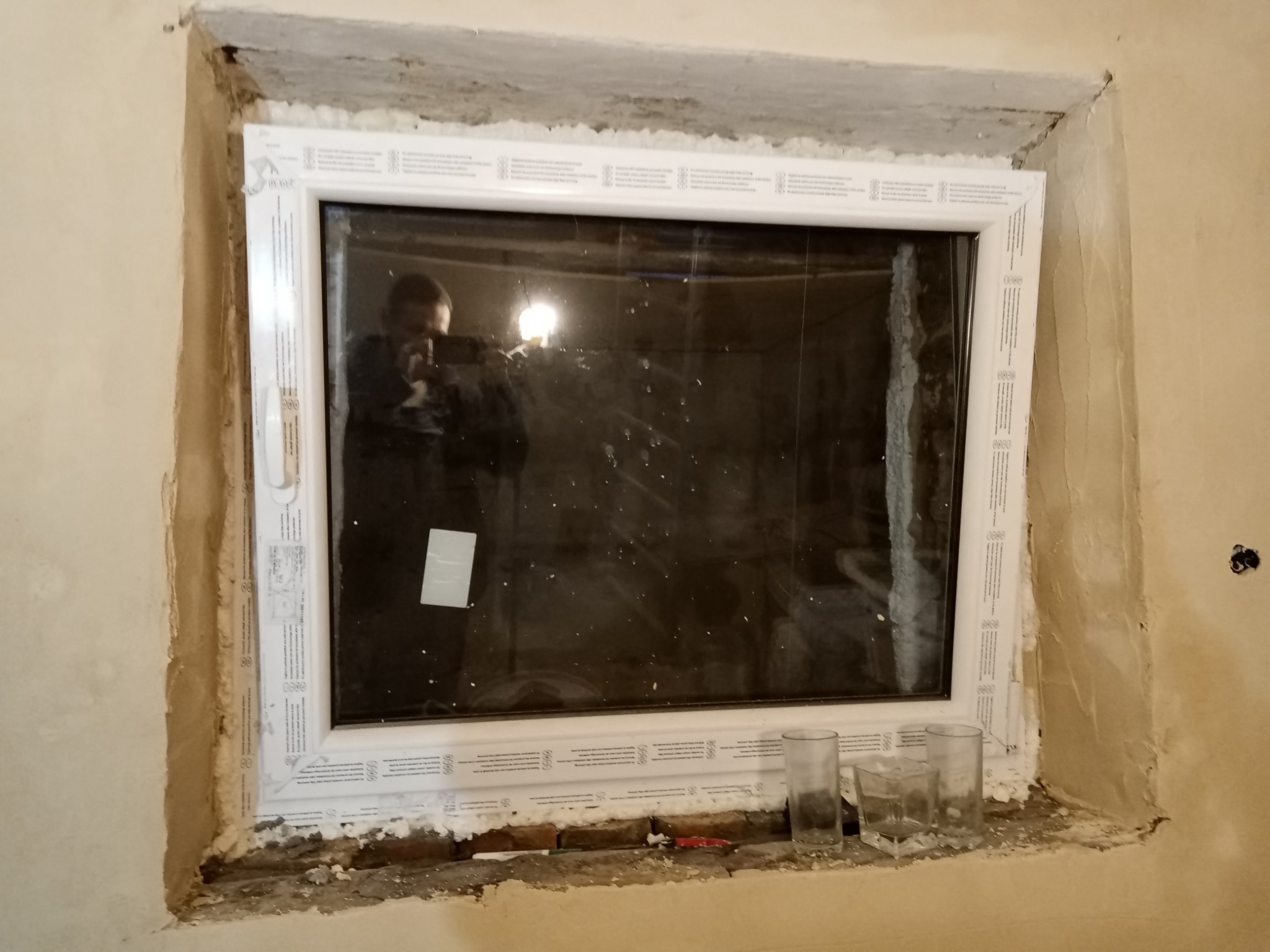 Nowe białe okno PCV zamontowane w surowym otworze okiennym, widoczne ślady pianki montażowej i niedokończone obróbki murarskie. W szybie odbija się osoba robiąca zdjęcie.
