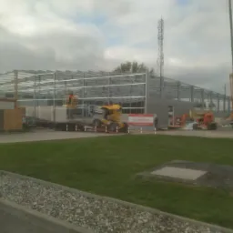 Montaż stalowej konstrukcji hali produkcyjnej, widoczny szkielet budynku, żuraw budowlany i podnośnik koszowy na tle pochmurnego nieba.